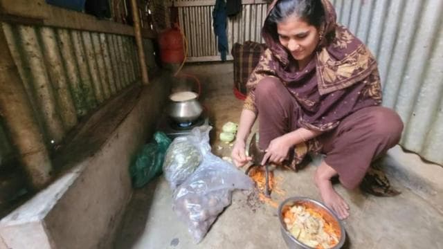 টিনের বেড়া দেওয়া ঘরে একটি দা নিয়ে সবজি কাটছেন একজন নারী