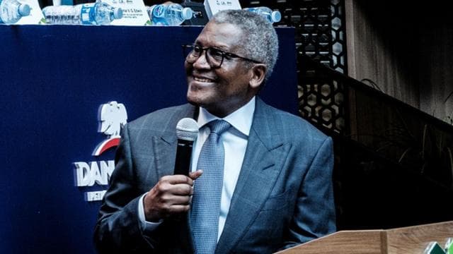 স্যুট পরা অ্যালিকো ড্যাঙ্গোটে ছবির বাম দিকে তাকিয়ে হাসছেন। তার ডান হাতে একটি মাইক্রোফোন রয়েছে।