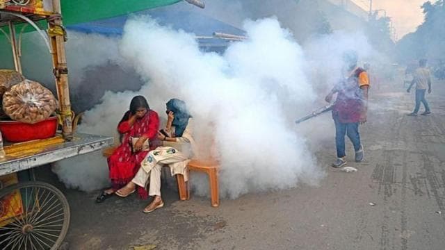 মশা নিয়ন্ত্রণে ফগার মেশিন দিয়ে ধোঁয়া ছাড়ছেন একজন কর্মী, সামনে একটি ভাসমান খাবারের দোকানের পাশে একটি বেঞ্চে বসে আছেন দুইজন নারী